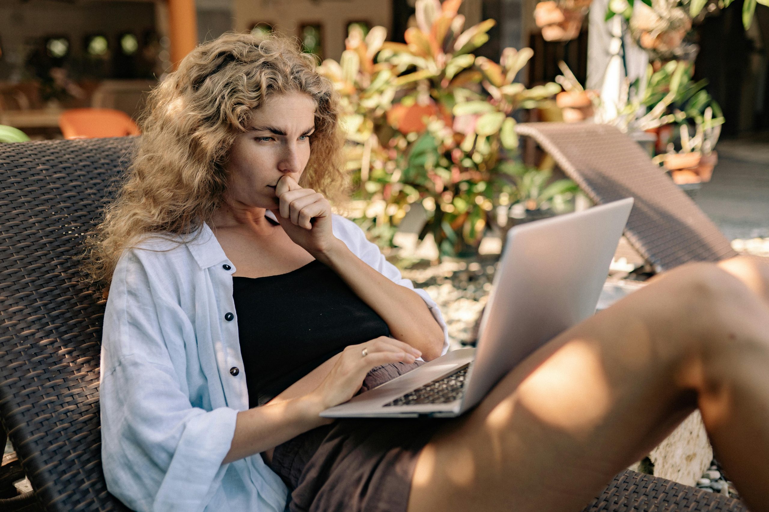 Woman with curly hair working remotely on a laptop while lounging outdoors in a tropical setting.